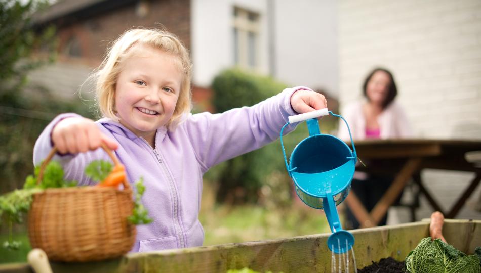 A little girl in a purple hoodie, stood in a garden holding food in a basket and a watering can.