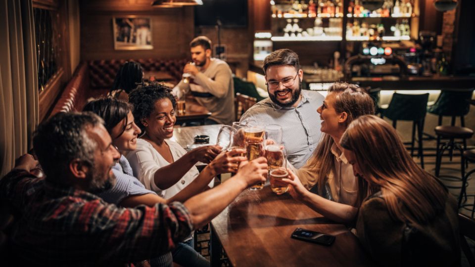 A photograph of a pub in England. There are people sat round a wooden table with pints of beer, lager and cider. They are smiling laughing and socialising.