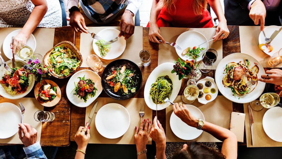 A photograph of a top down view of a table in a restaurant with food on the plates. You can see lots of hands using cutlery and utensils to grab specific food.