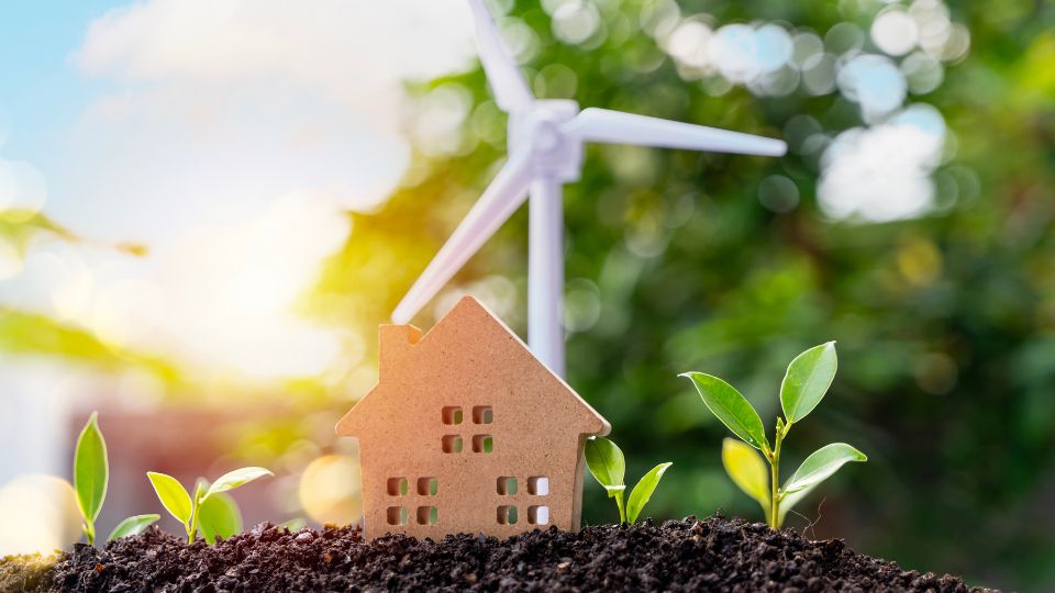 Cardboard cut out house in soil with a wind turbine in the background.